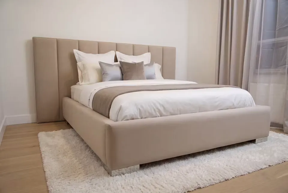 Full angled view of a beige upholstered bed with a low channel headboard and pale textured rug.