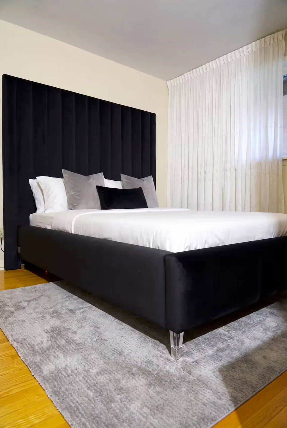 Black upholstered bed with a tall channel headboard and small metallic legs beside sheer curtains