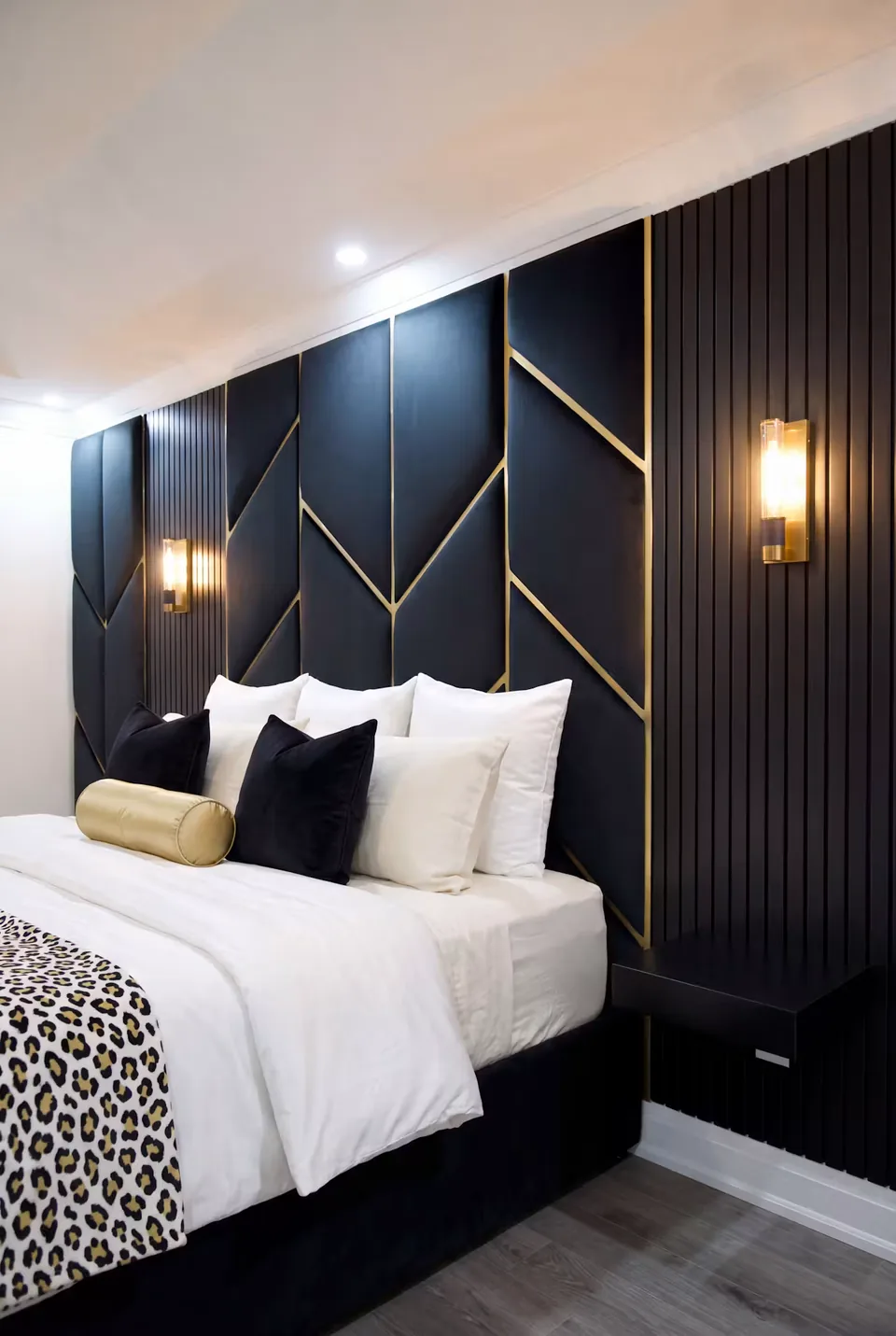 Right angled view of the black bed and gold chevrons between warm wall lights.