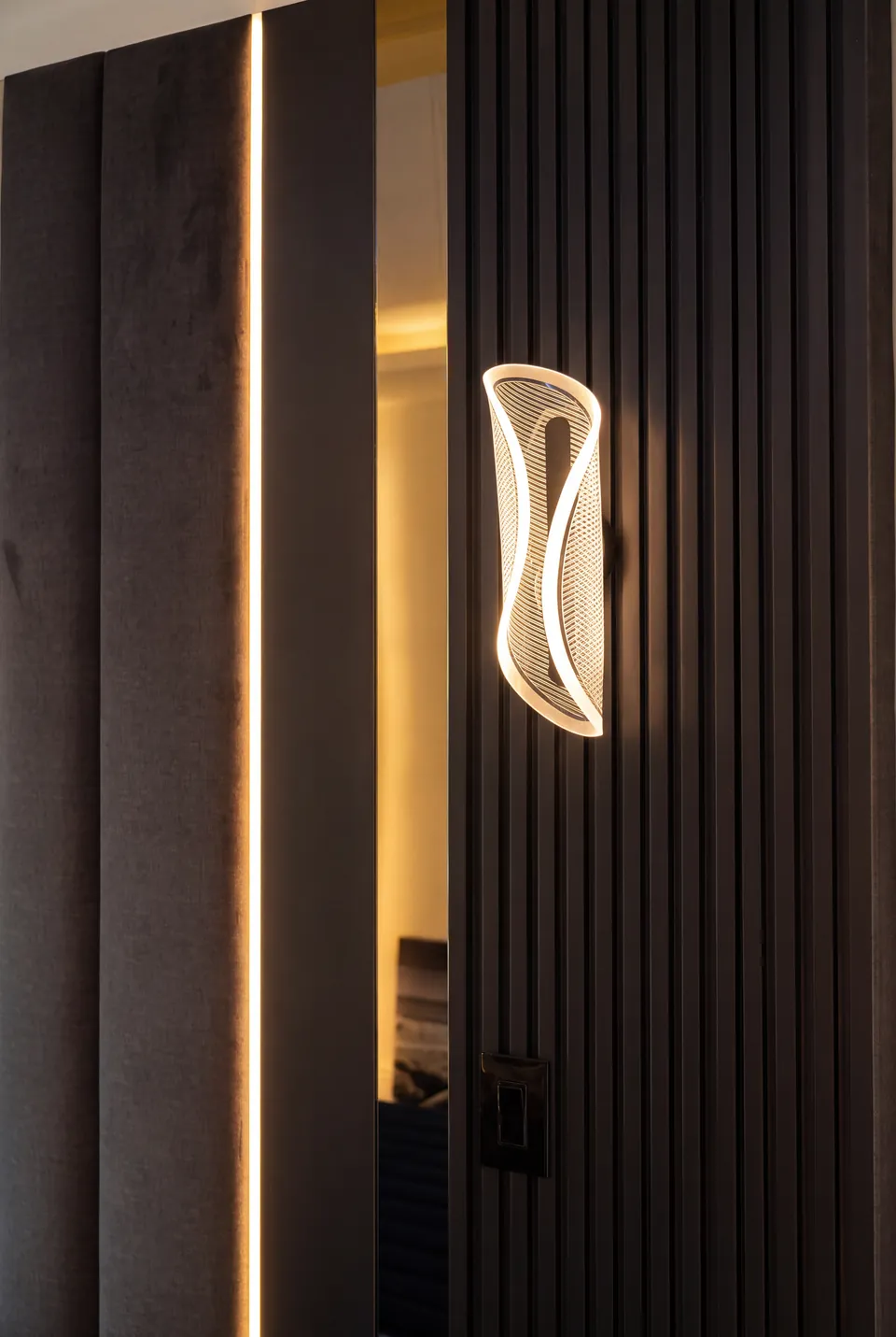 Detail of a curved wall sconce beside dark slats, a mirror and a warm illuminated panel edge.