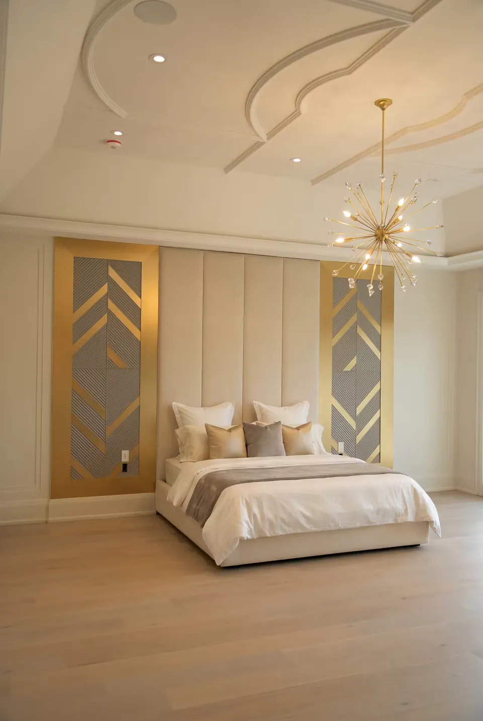 Wide angled view of the cream bed and gold geometric wall accents beneath a starburst chandelier.