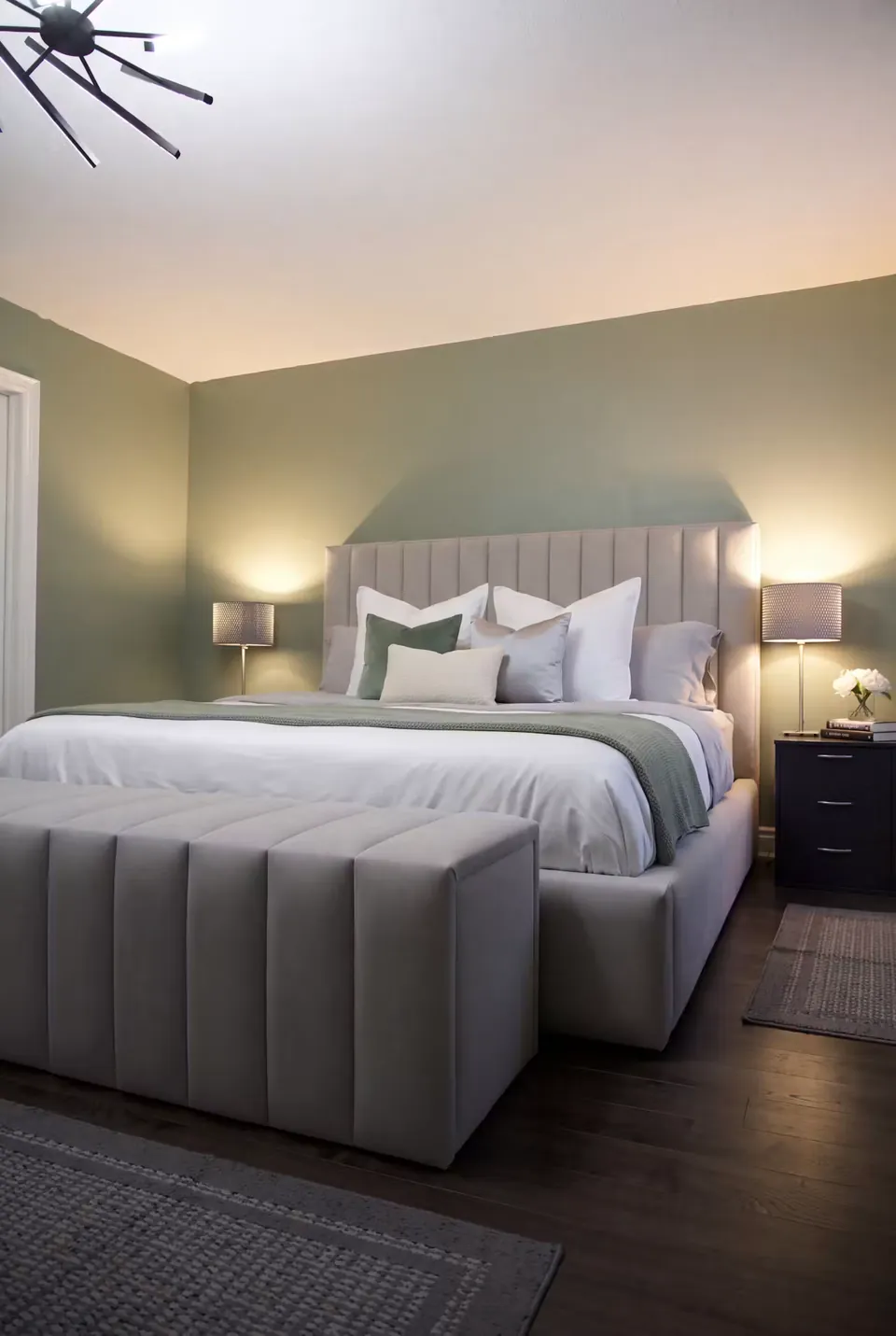 Angled view of the grey upholstered bed and channelled bench beside a warm bedside lamp.