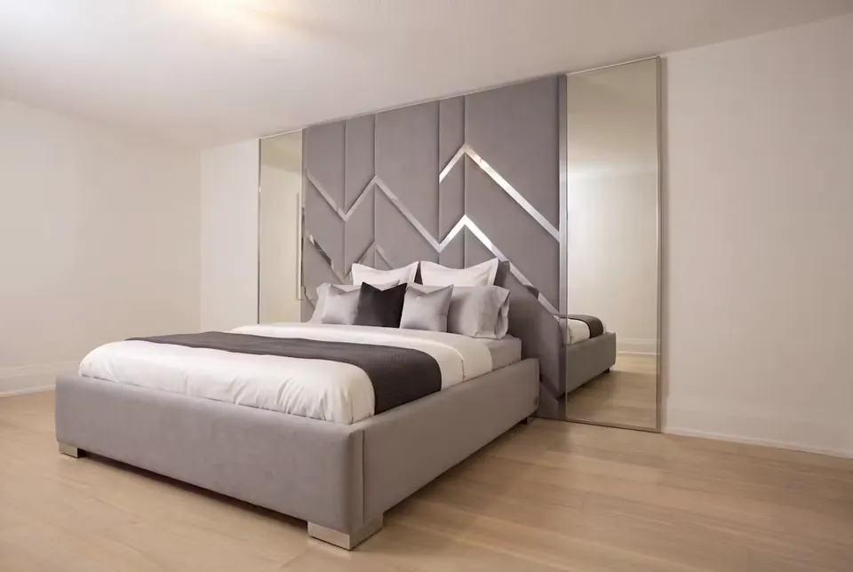 Full angled view of a grey upholstered bed beneath gold-accented chevron wall panels and mirrors.