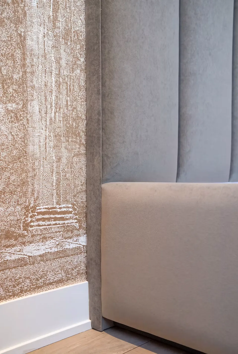 Detail of a lower grey upholstered headboard overlapping tall padded channels beside patterned wallpaper.