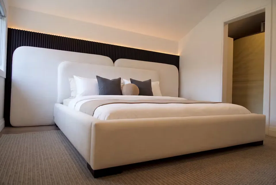 Angled view of the ivory bed beside a window and a black upholstered wall backdrop