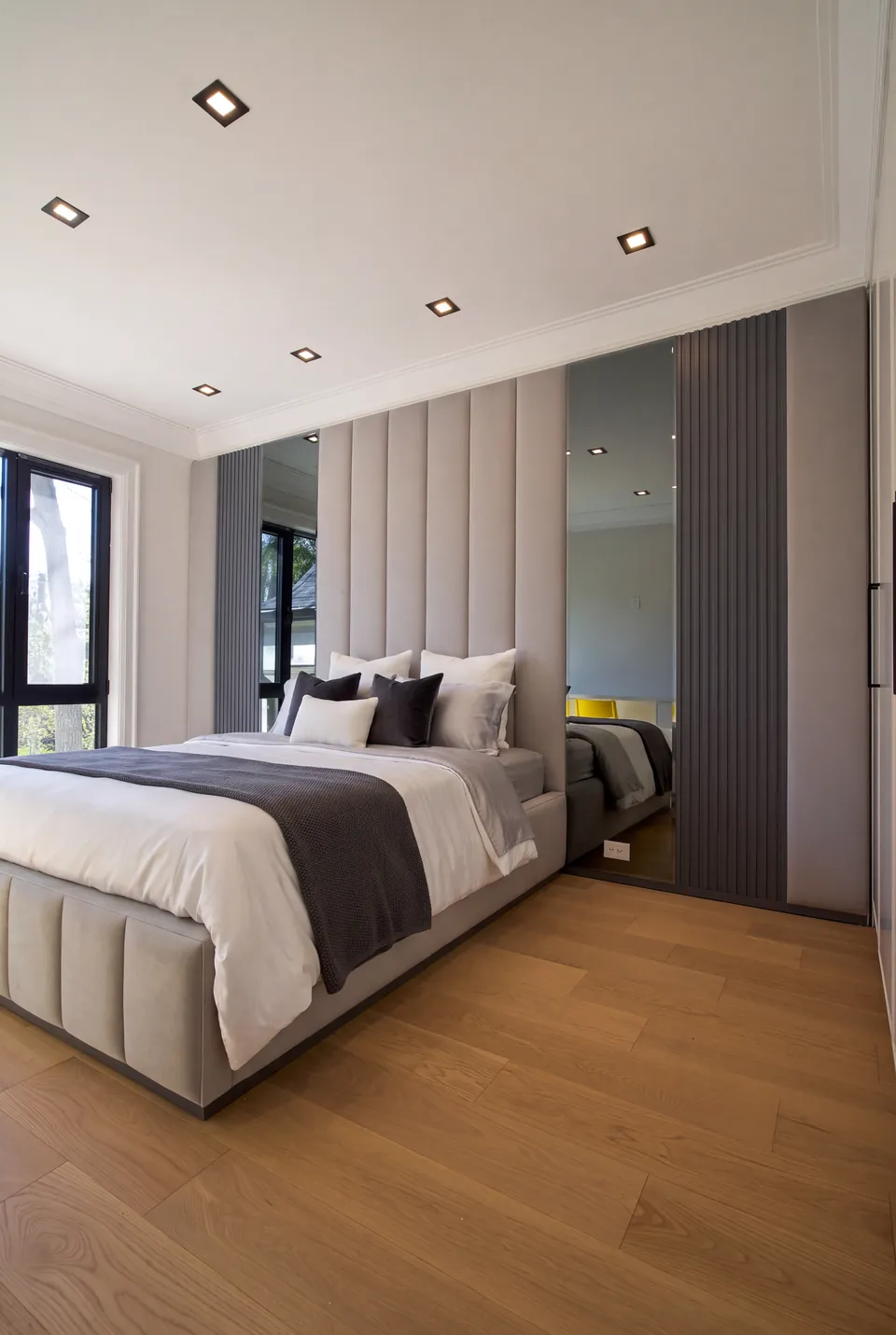 Wide angled view of a pale upholstered bed beneath tall channels flanked by mirrors and dark slats.