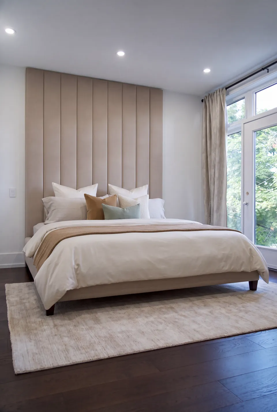 Full view of a sand-coloured upholstered bed with tall vertical wall panels beside large windows.