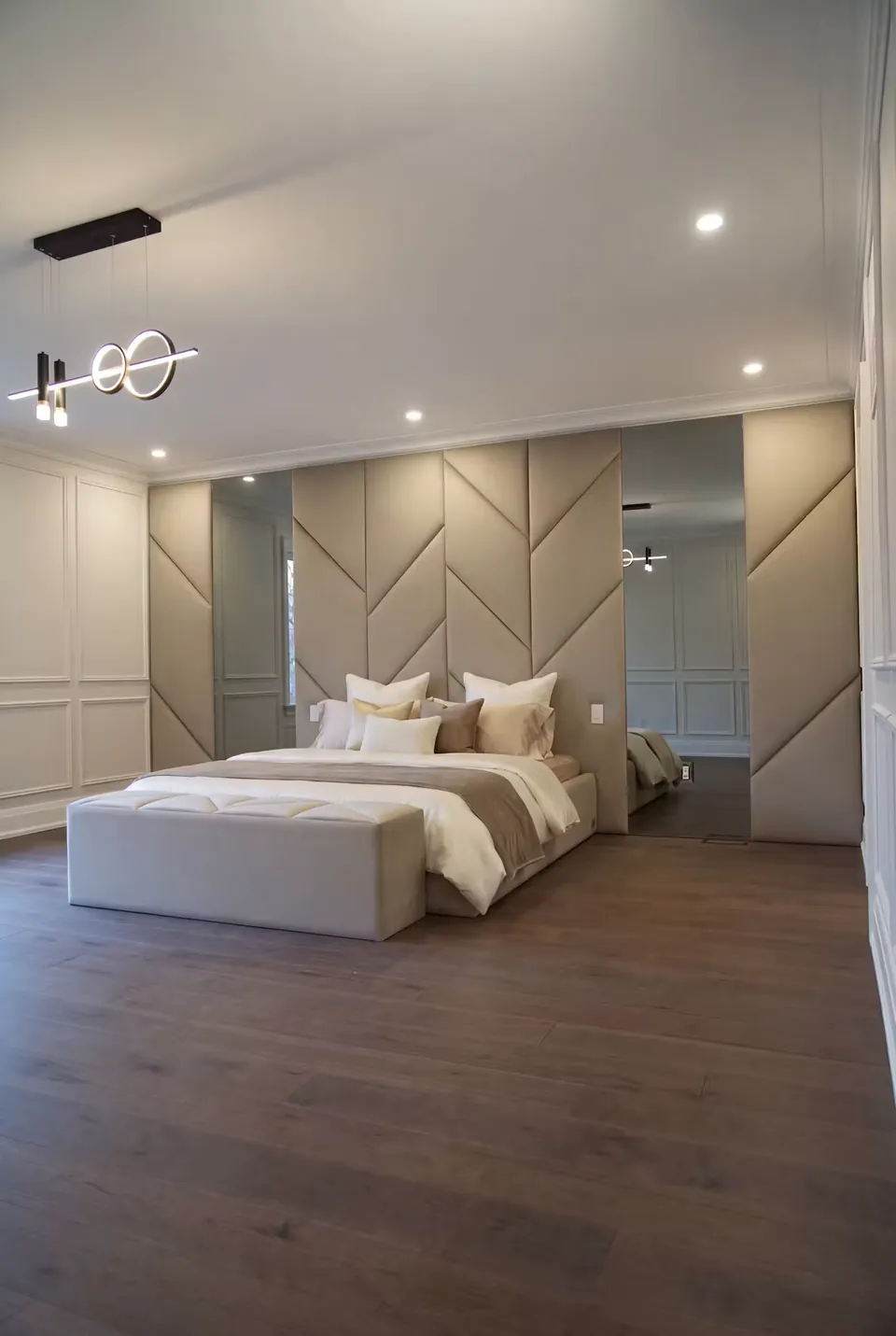 Full angled view of a taupe upholstered bed beneath geometric panels and mirrored wall strips.