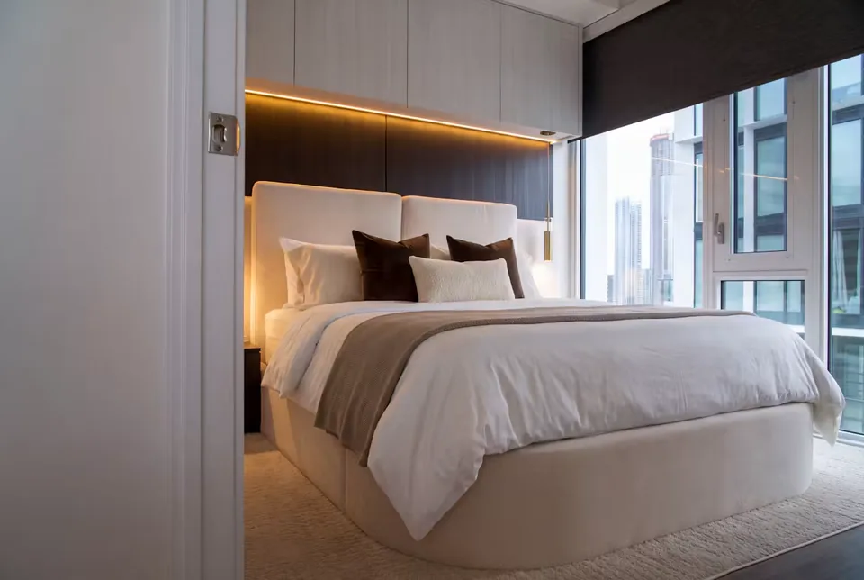 Full angled view of a taupe bed beneath pale vertical panels and a dark headboard with amber lighting.