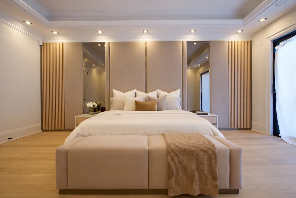 Full front view of a taupe bed and matching bench beneath mirrored and slatted wall panels.