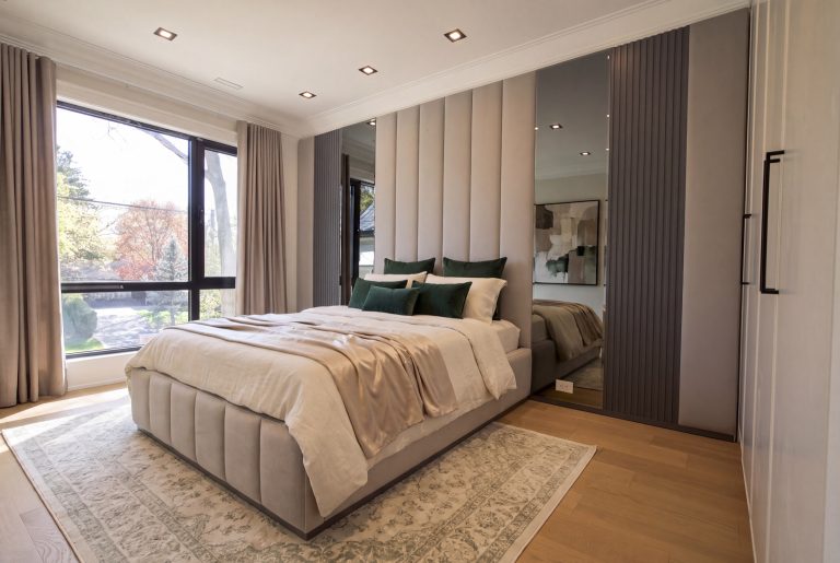 Beige upholstered bed with a tall paneled headboard, mirrored wall panels and green accent pillows by BOKO Designs.