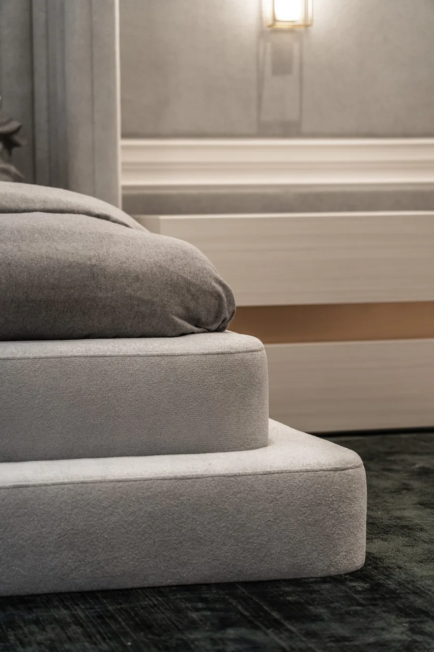 Rounded grey upholstered bed base with two layered edges beside a bedside table