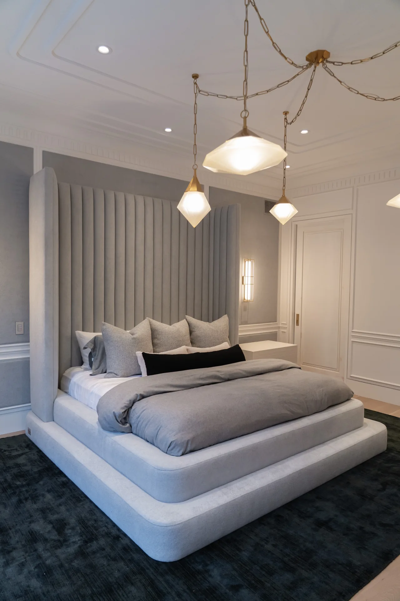 Grey upholstered bed with a tall channelled headboard, layered base and pendant lights